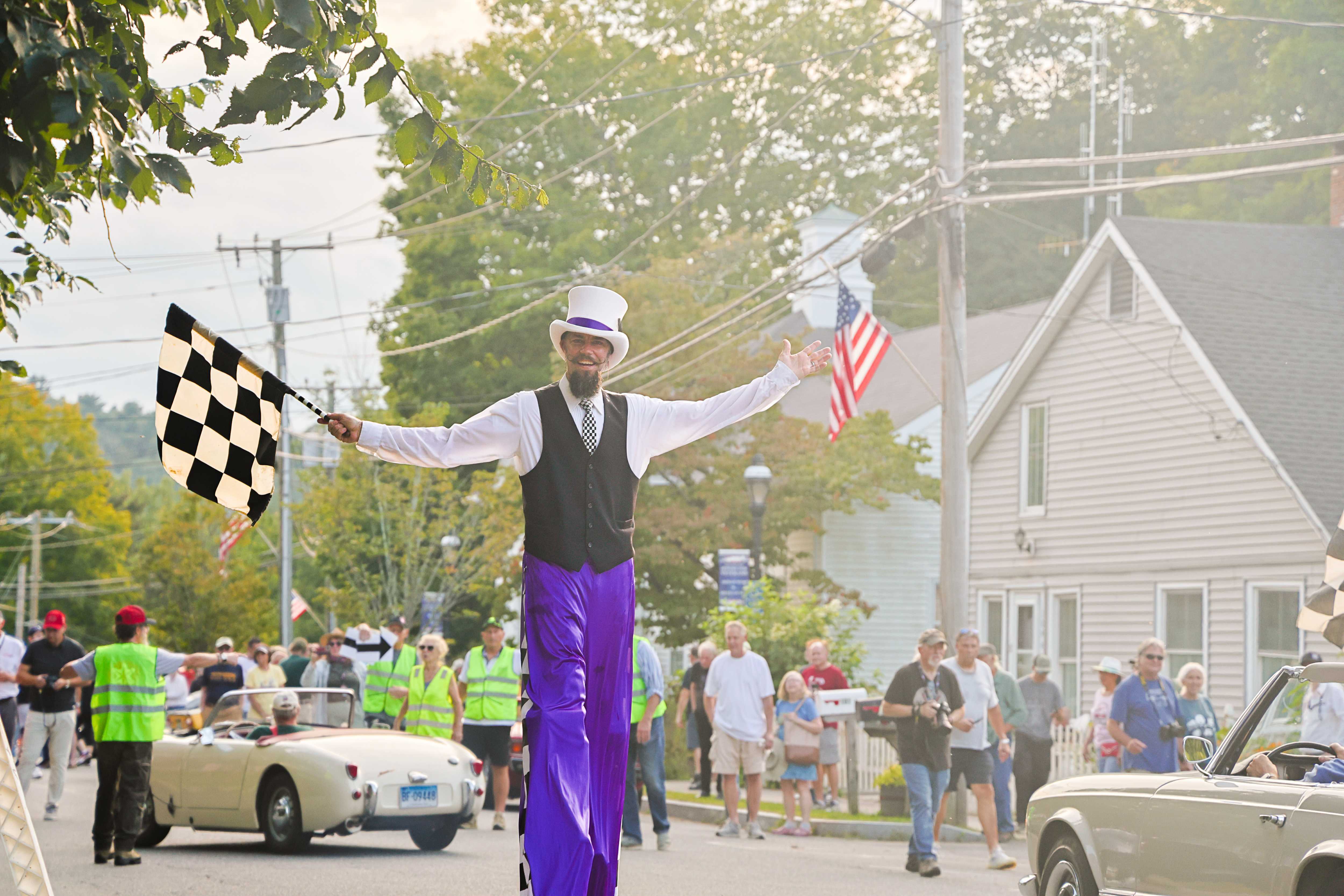 A smiling stilt walker holds a checkered flag with arms outstretched, welcoming cars into Falls Village during the parade.