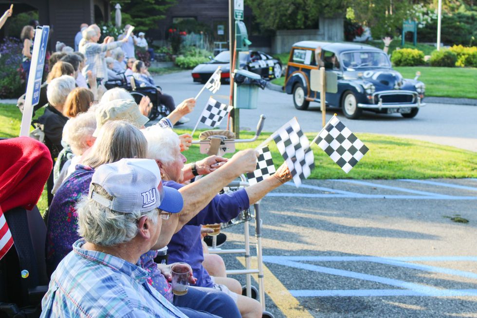 Elderly spectators wave checkered flags as classic cars pass by during the parade.